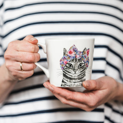 This white china mug features an illustration of a cat by renowned artists Tipperleyhill. A black and white sketch of head and neck of a tabby cat with a pretty colourful and flowery scarf tied around its head, finished in a bow.