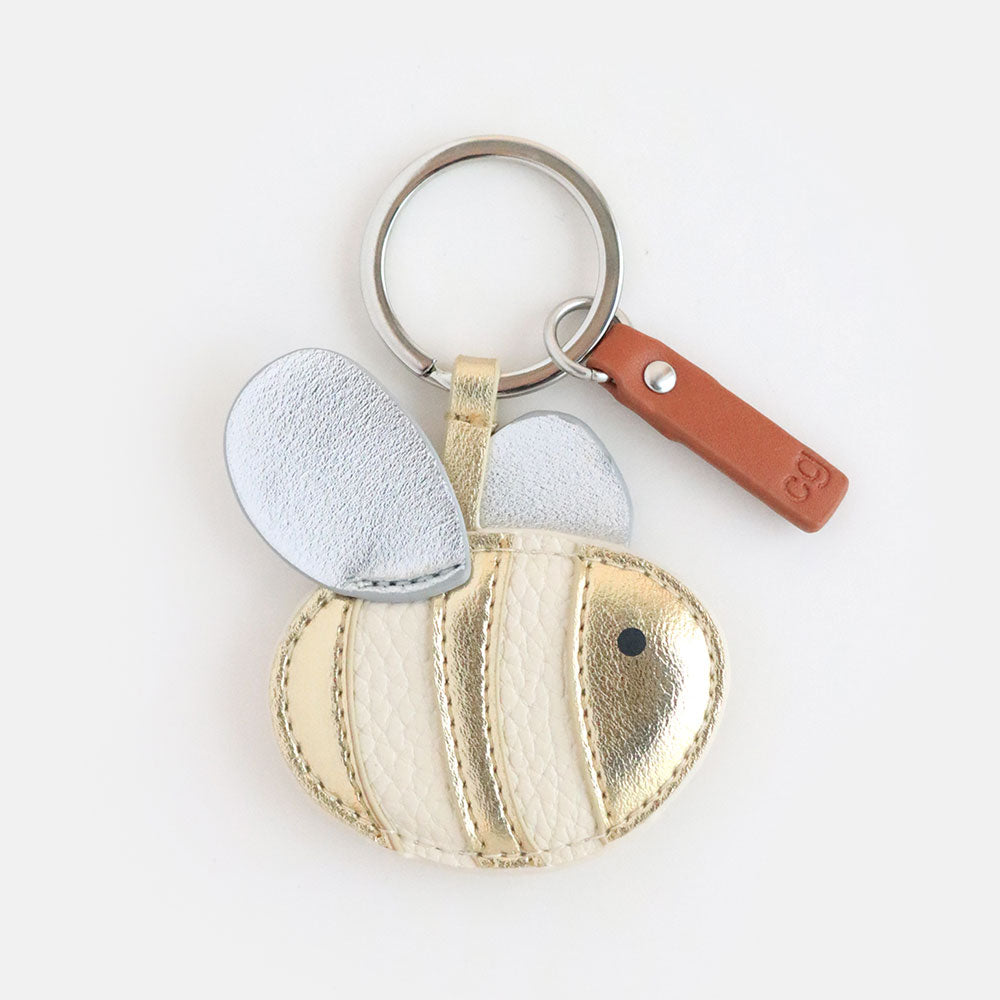 This novelty keyring from Vinegar Hill features a bee with an egg shaped body in cream vegan leather that is appliqued with stripes of gold leather look fabric. It has a printed black dot for an eye and silver ovoid shapes stitched on front and back for wings. The bee is attached to the silver coloured ring with a strip of the gold fabric and completed with a tan leather look brand tag.  