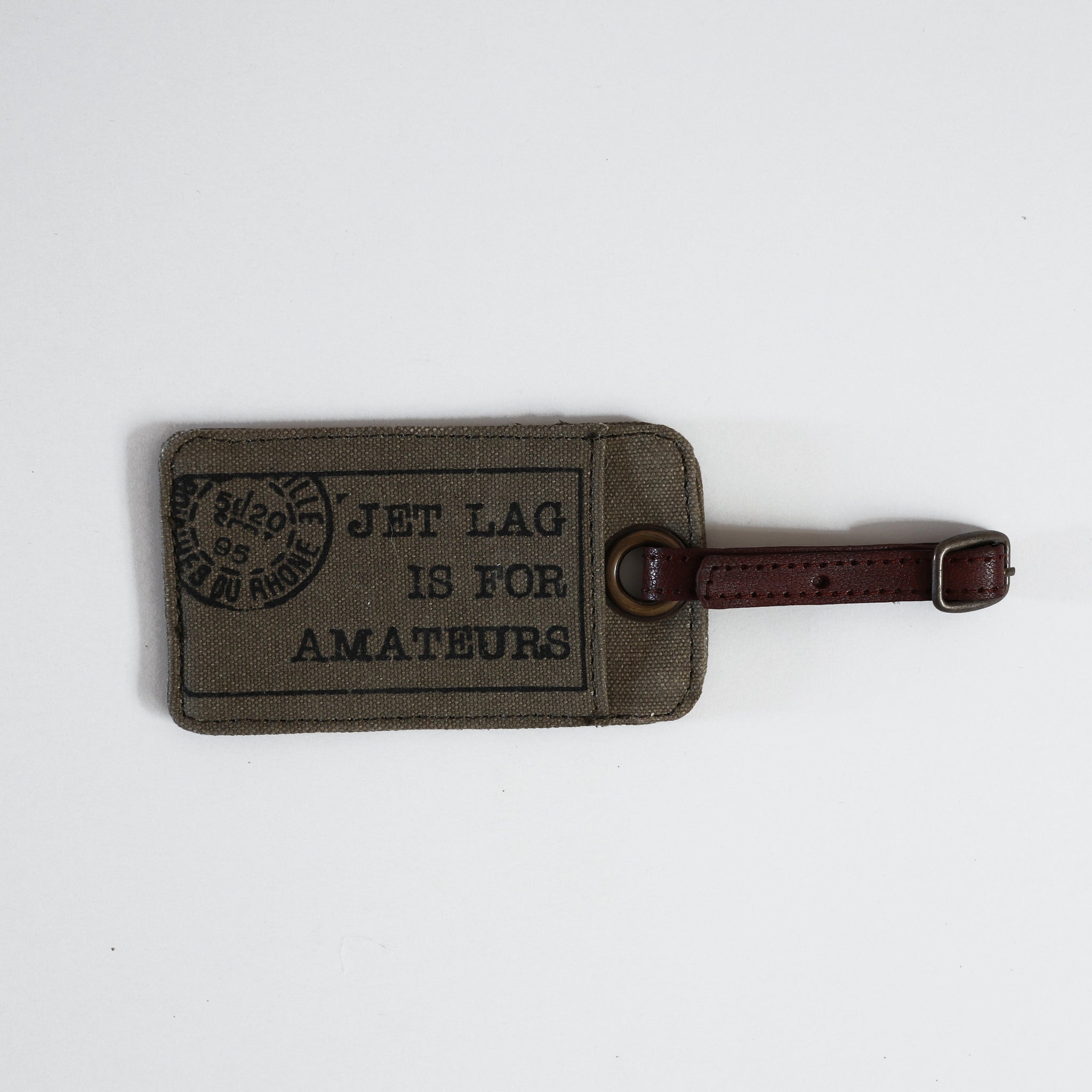 Brown luggage label, from Vinegar Hill. The main body of the label has ‘JET LAG IS FOR AMATEURS’ printed in a black serif typeface. A strap in vegan leather with a buckle fastening is attached with a metal eyelet. 
