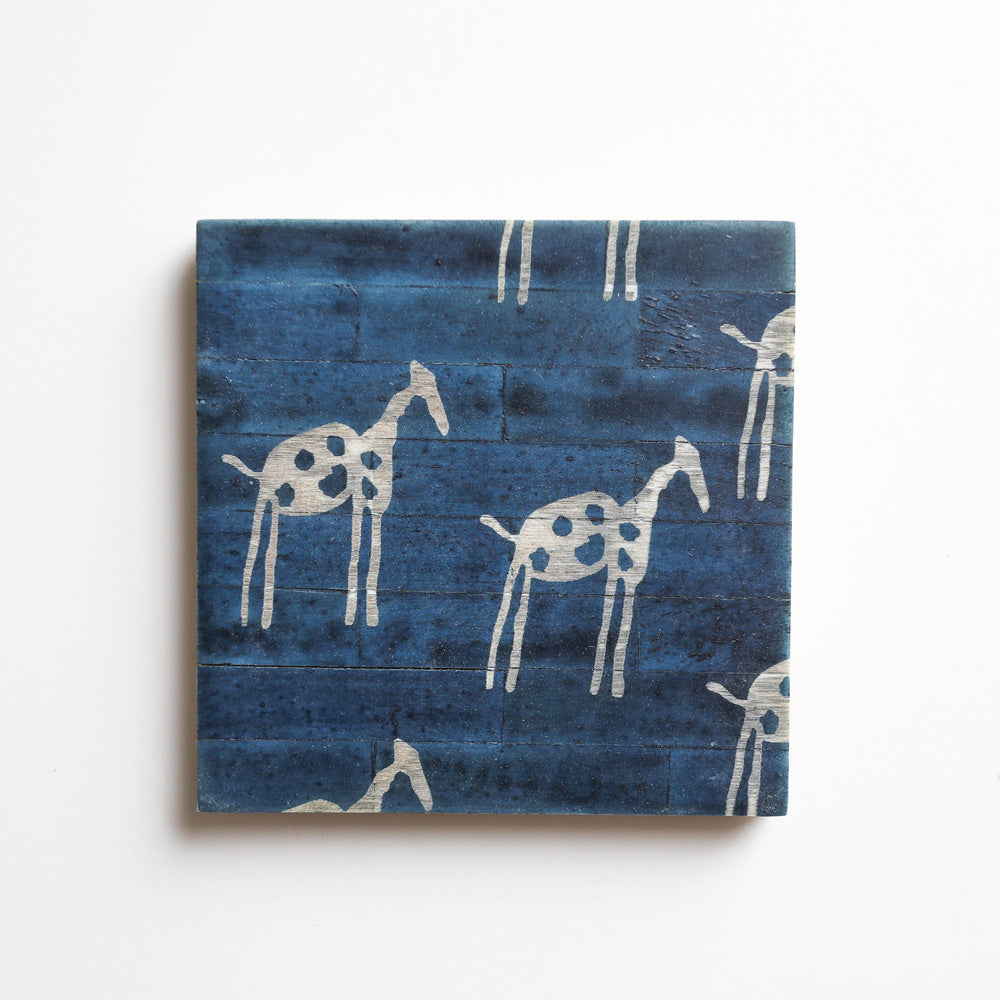 A square coaster from Vinegar Hill. The image shows a coaster with a mid blue background and a haphazard primitive ‘cave art’ look showing giraffes in white – shown side on walking left to right.