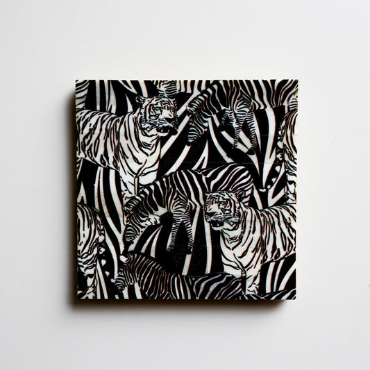 A square coaster from Vinegar Hill. The image shows a coaster with a black background and tigers and zebras in white – a lino cut appearance – they are in different poses, amongst long grass.