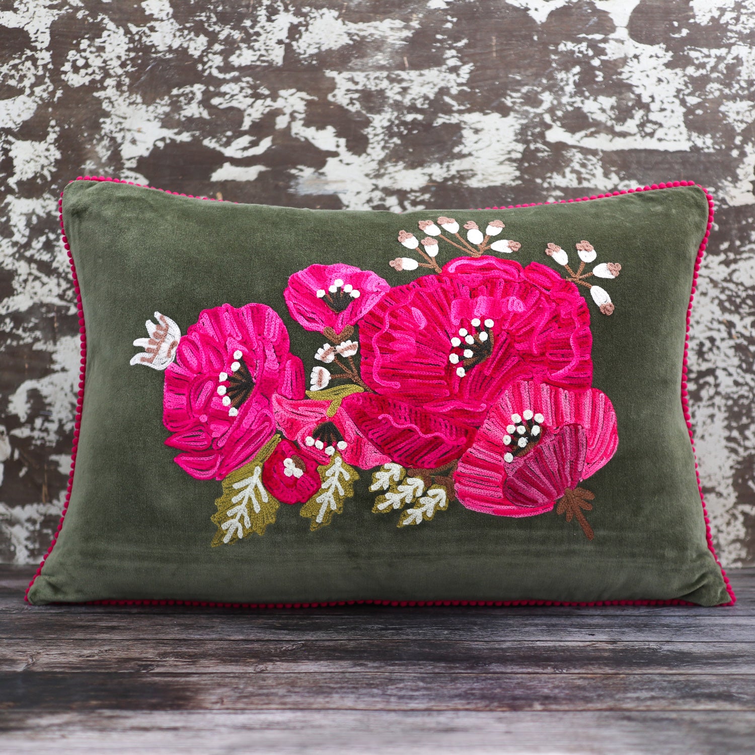 A Vinegar Hill own brand cushion is shown, an olive green velvet rectangular cushion has intricate embroidery centred over majority of the front – three large blooms in shades of pink dominate, with smaller blooms in the same shades of bright pink and sprigs in cream and pale brown. There is foliage in green and cream and stems and stamens in shades of brown.