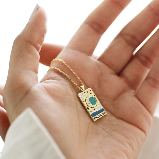 A gold plated pendant with enamel detail on a gold plated trace chain by Lisa Angel at Vinegar Hill. The pendant is inspired by a traditional tarot card and depicts a crescent moon in turquoise enamel infill, with tiny black enamel stars, white rolling clouds and a stretch of blue sea. Beneath the image is 'the moon' written in bold black lettering. Shown being held displayed across the palm of a model’s hand.