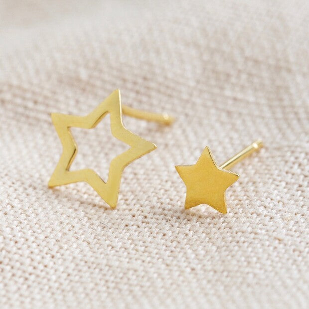 A close up of a pair of star stud earrings by Lisa Angel from Vinegar Hill. The left stud is the larger and is the outline of the star, the other, on the right, is the solid cut out of the first. Shown on a cream fabric background.