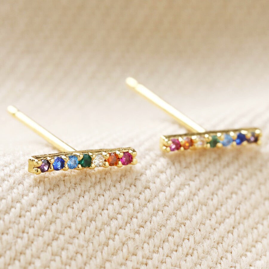 A pair of gold plated multi coloured crystal bar ear studs by Lisa Angel from Vinegar Hill. The narrow dainty bars have seven different coloured crystals inset. They are - amethyst purple, sapphire blue, aqua marine blue, emerald green, diamante, citrine orange and ruby red.  The bars are in a horizontal position and are shown in close up on a cream fabric background. 
