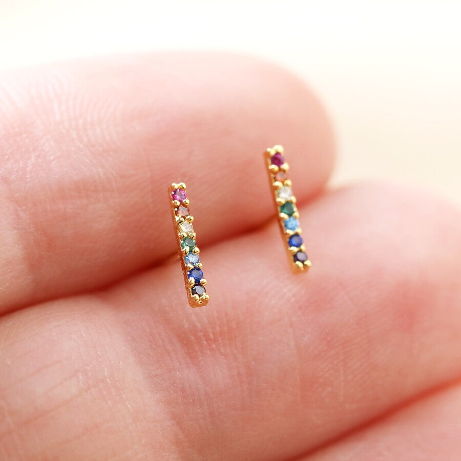 A pair of gold plated multi coloured crystal bar ear studs by Lisa Angel from Vinegar Hill. Shown being held, vertically, between two fingers, the narrow dainty bars have seven different coloured crystals inset. From bottom to top they are - amethyst purple, sapphire blue, aqua marine blue, emerald green, diamante, citrine orange and ruby red. 