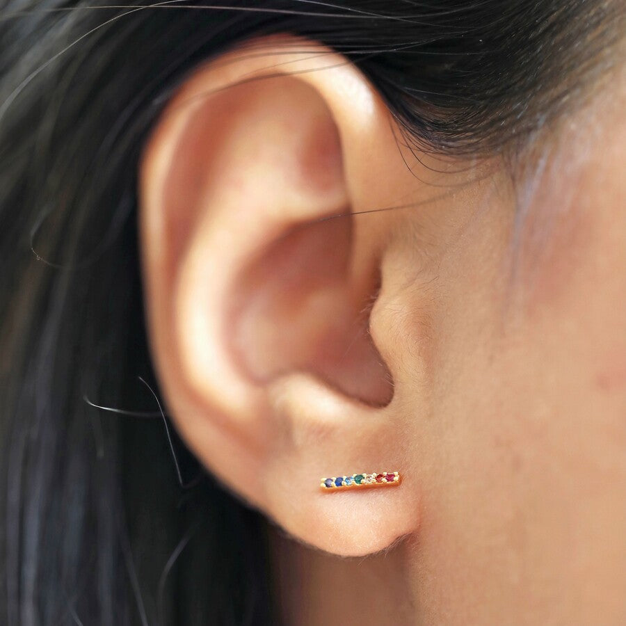 A gold plated multi coloured crystal bar ear stud by Lisa Angel from Vinegar Hill. Shown being worn horizontally in a model’s ear lobe. From left to right they are - amethyst purple, sapphire blue, aqua marine blue, emerald green, diamante, citrine orange and ruby red. 